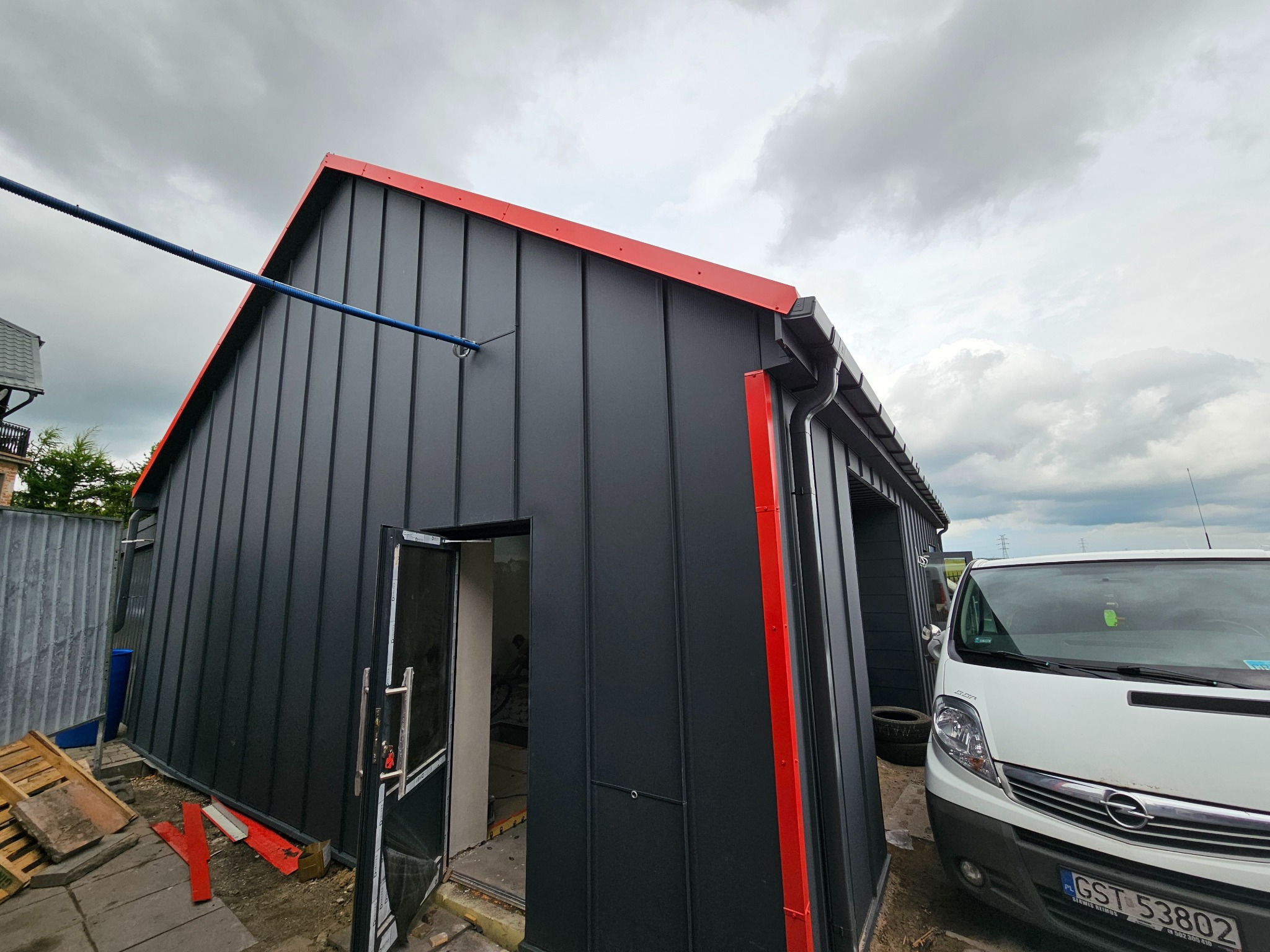 Nowoczesny, czarny budynek z blachy trapezowej, wykończony czerwoną obróbką. Widoczne rynny i częściowo otwarte drzwi. Na pierwszym planie biały samochód dostawczy.