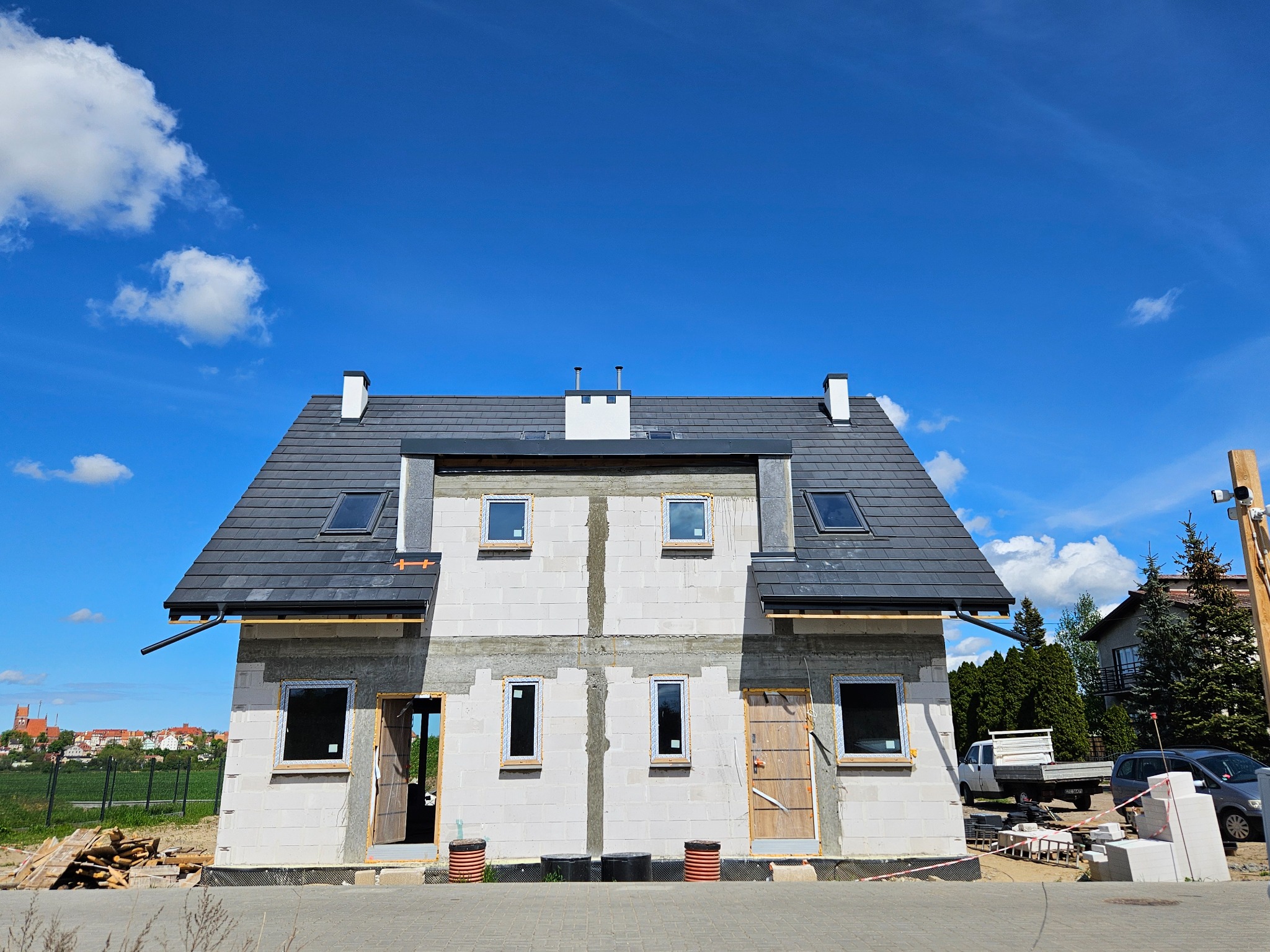 Dom w stanie surowym zamkniętym z ciemną dachówką i oknami, widok z zewnątrz w słoneczny dzień. W tle zabudowa miejska i błękitne niebo z chmurami.