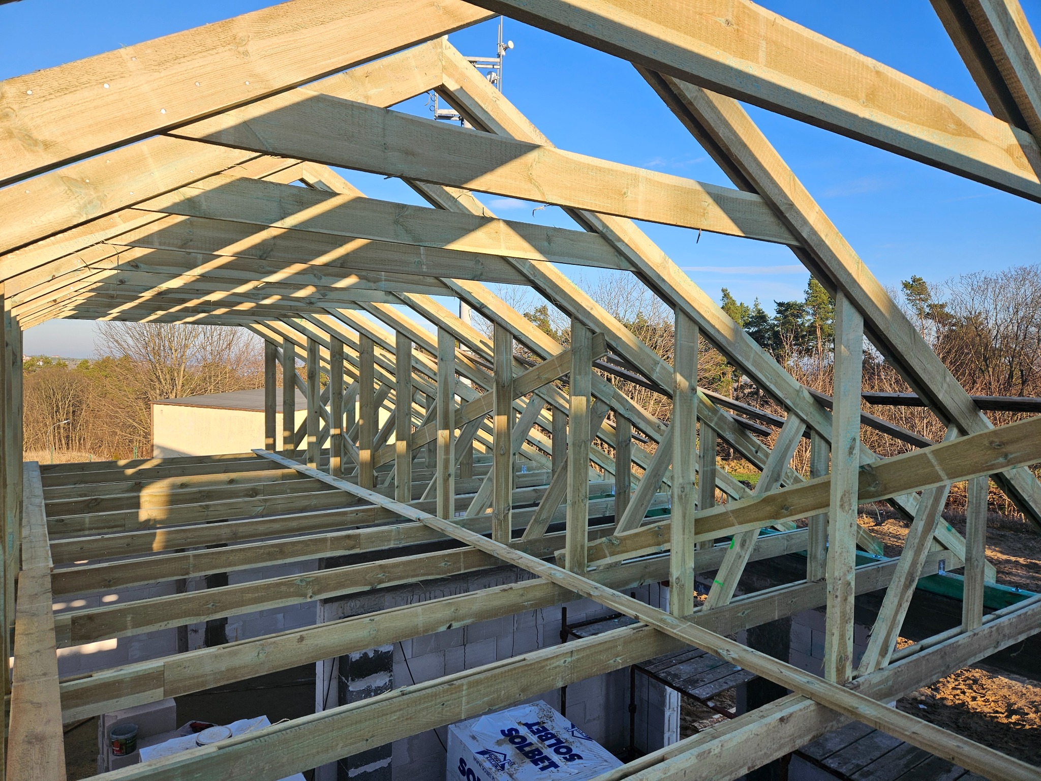 Konstrukcja drewniana dachu w trakcie budowy, widok z wnętrza. Elementy więźby dachowej z jasnego drewna na tle błękitnego nieba i drzew.
