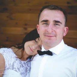 Portret uśmiechniętego pana młodego w białej koszuli i czarnej muszce, z panną młodą opierającą się o jego ramię w koronkowej sukni ślubnej, na tle drewnianej ściany.