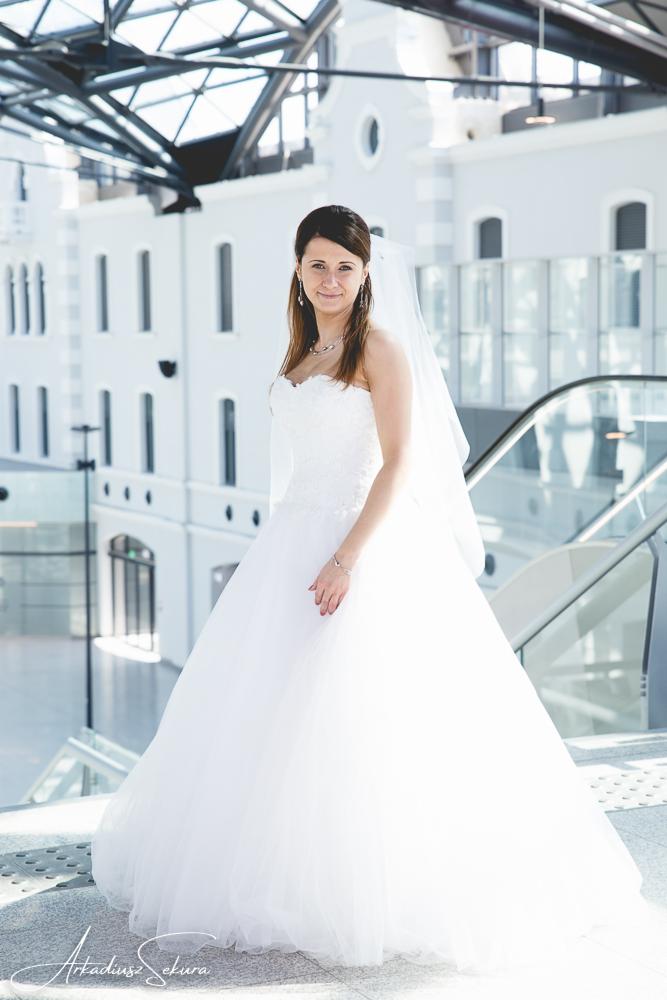Panna młoda w białej sukni balowej i welonie, pozuje na tle nowoczesnego budynku z przeszklonym dachem i schodami ruchomymi.