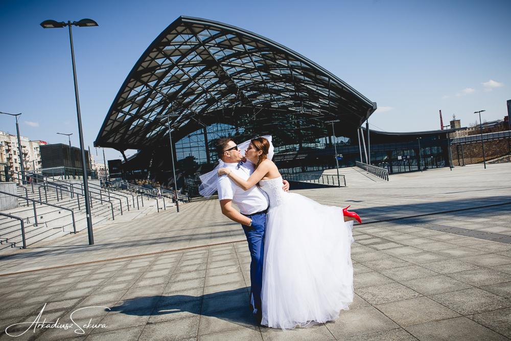 Para młoda w objęciach, panna młoda w białej sukni i czerwonych szpilkach, pan młody w białej koszuli i niebieskich spodniach na tle nowoczesnego dworca kolejowego w słoneczny dzień.
