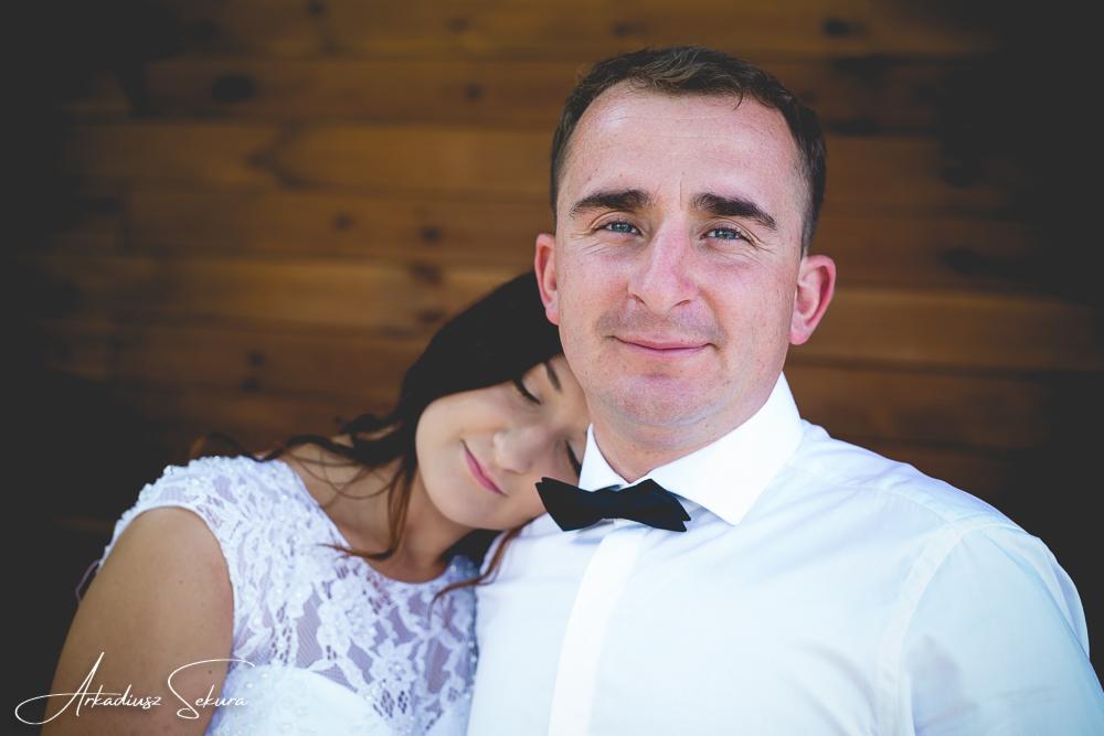 Portret uśmiechniętego pana młodego w białej koszuli i czarnej muszce, z panną młodą opierającą się o jego ramię w koronkowej sukni ślubnej, na tle drewnianej ściany.