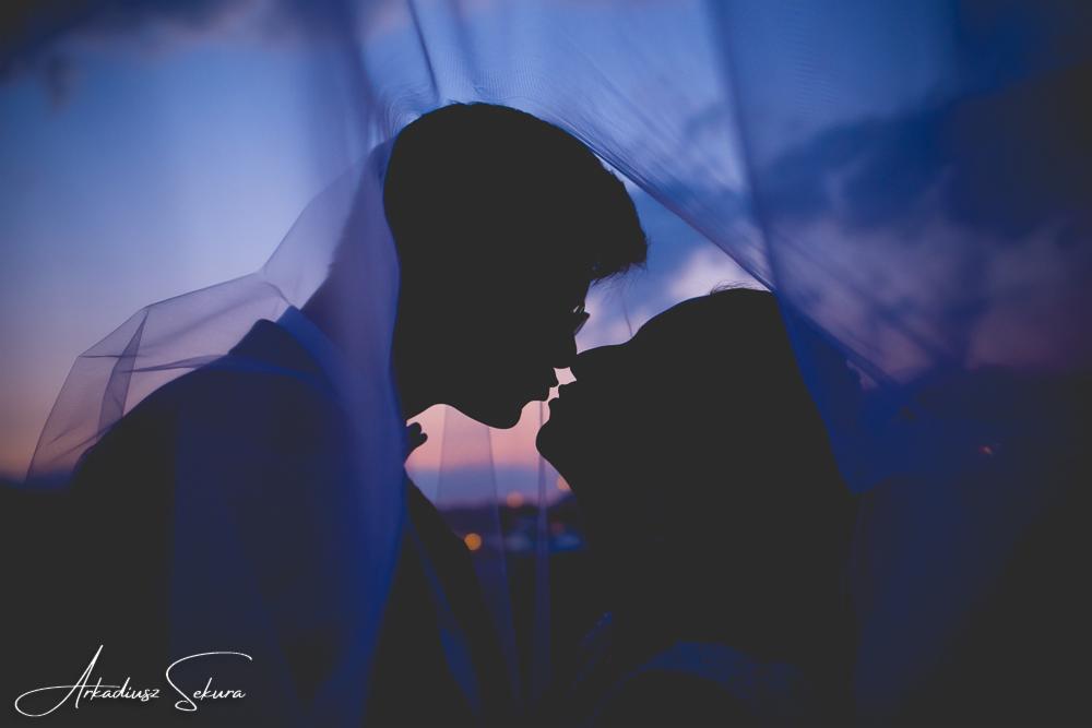 Silhouettes of a kissing couple framed by a flowing veil against a sunset sky, with the photographer's signature visible at the bottom.
