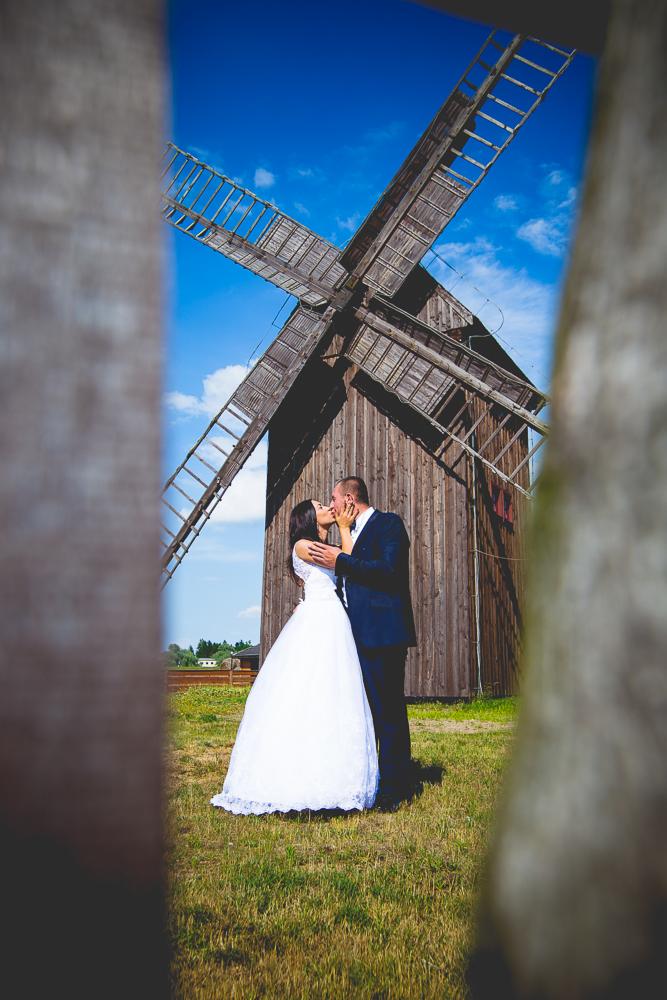 Para młoda całuje się na tle drewnianego wiatraka, widziana przez fragment drewnianej konstrukcji w pierwszym planie.