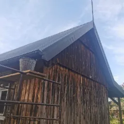 Metalowy dach w kolorze grafitowym na drewnianym budynku gospodarczym w Brąszewicach, widok z poziomu rusztowania z wiadrem. Nowe orynnowanie.