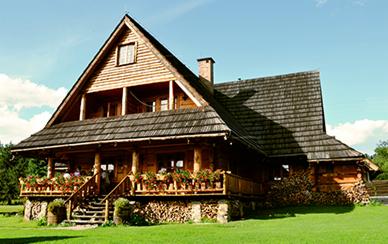 Drewniany dom w stylu góralskim z werandą, kwiatami i stertą drewna na trawniku w słoneczny dzień.