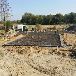 Przygotowanie zbrojenia pod płytę fundamentową na budowie, widoczny biały van i pracownicy w tle.