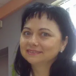 Portret uśmiechniętej kobiety z ciemnymi włosami i delikatną biżuterią na tle biura.