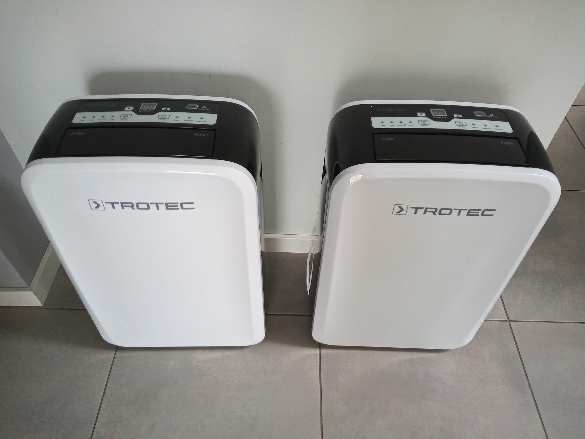 Dwa białe osuszacze powietrza marki Trotec z czarnym panelem sterowania, ustawione obok siebie na szarej podłodze z płytek.