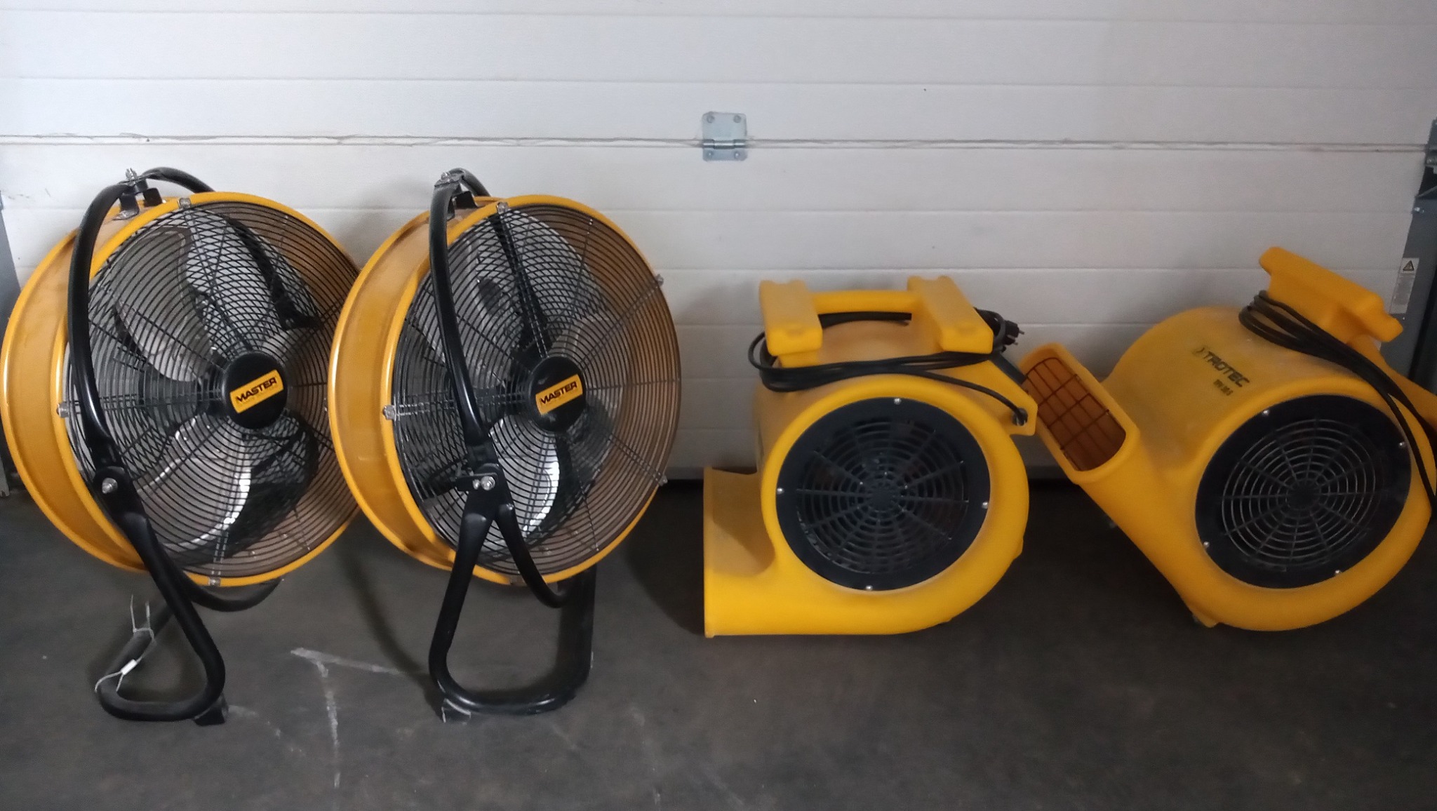 Dwa wentylatory osuszające Master i dwa dmuchawy Trotec ustawione obok siebie na betonowej podłodze, na tle białej bramy garażowej.