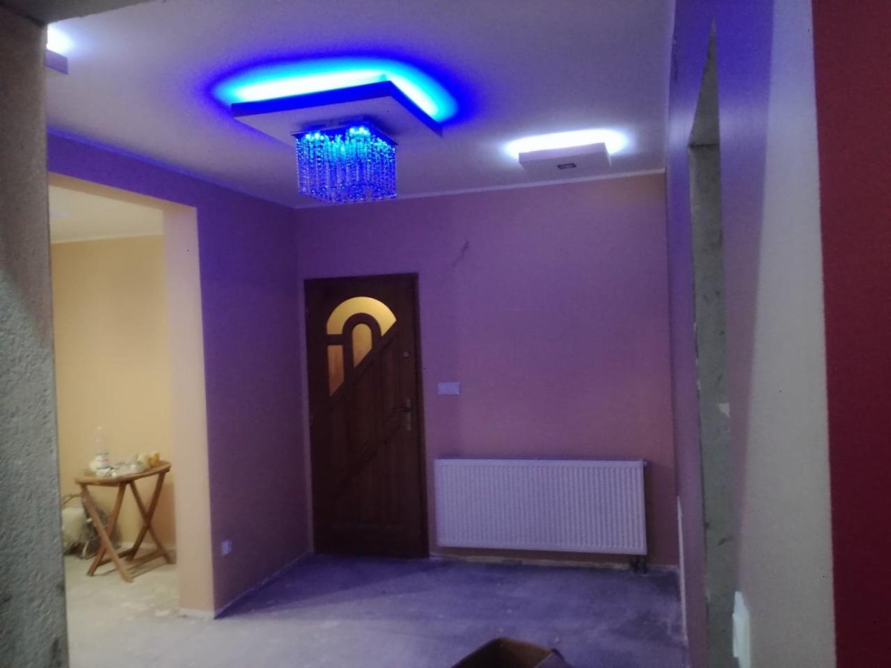 Nowoczesne wnętrze po remoncie: ściany w odcieniach fioletu, biały grzejnik, drewniane drzwi z dekoracyjnym wycięciem, wielobarwne oświetlenie sufitowe z elementami kryształowymi. Widoczny fragment...