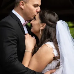 Panna młoda w białej sukni i welonie, obejmowana przez pana młodego w czarnym garniturze z różową muchą, który całuje ją w czoło.