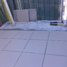 Układanie jasnych płytek na balkonie z użyciem krzyżyków dystansowych, widoczne narzędzia: poziomica, młotek gumowy, młotek.