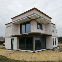 Dwukondygnacyjny dom murowany w stanie surowym otwartym, z widocznymi czarnymi ramami okiennymi i elementami betonowymi, otoczony gruzem i ziemią na niezagospodarowanej działce.