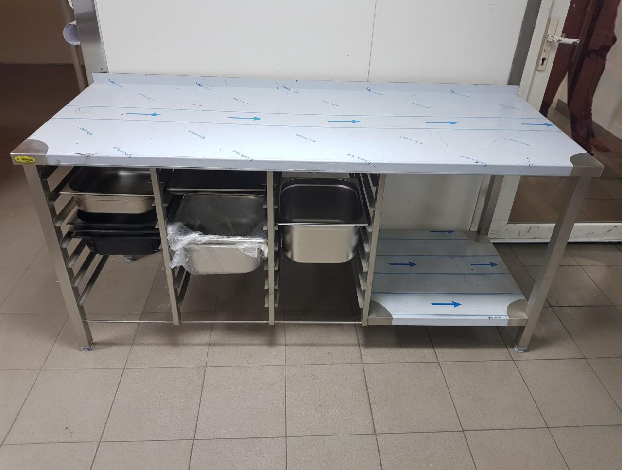 Stół roboczy ze stali nierdzewnej z pojemnikami gastronomicznymi, zabezpieczony folią ochronną, widoczny na tle kafelków podłogowych.