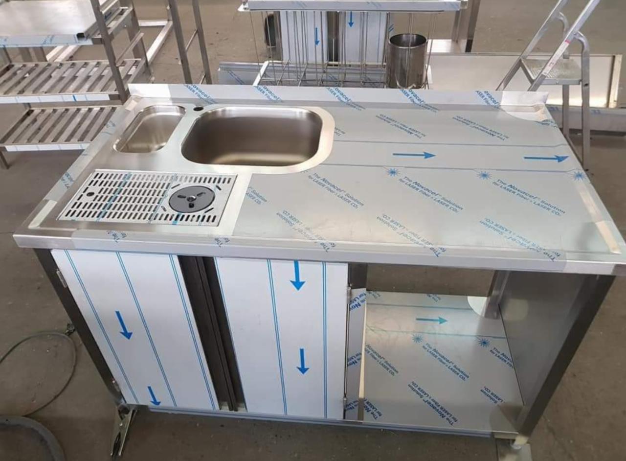 Stalowa konstrukcja barowa z zlewem i ociekaczem, pokryta folią ochronną z niebieskimi strzałkami, widoczne elementy wyposażenia zaplecza.