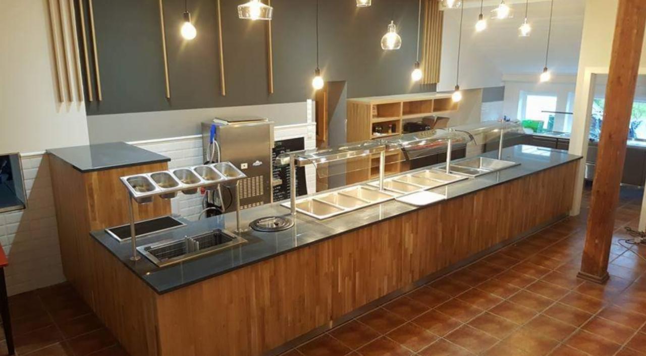 Długa lada barowa z drewnianymi panelami, czarnym blatem i metalowymi pojemnikami na składniki, oświetlona wiszącymi lampami, w lokalu z brązową podłogą i szaro-białymi ścianami.