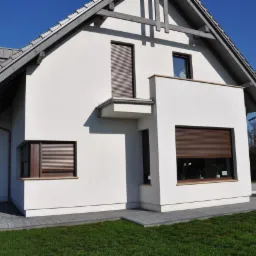 Dom jednorodzinny z białymi ścianami i brązowymi roletami zewnętrznymi, widok z zewnątrz na elewację z oknami zabezpieczonymi roletami w kolorze brązu.