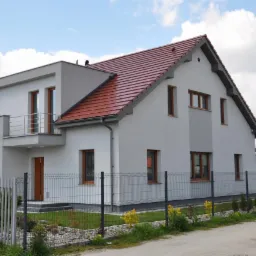 Nowoczesny dom jednorodzinny z szarą elewacją, czerwoną dach&oacute;wką i drewnianymi ramami okiennymi, otoczony metalowym ogrodzeniem panelowym i zielenią.