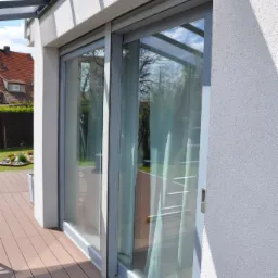 Przesuwne okno tarasowe z szarą ramą, widok z zewnątrz na drewnianą podłogę tarasu i dom z czerwonym dachem w tle.