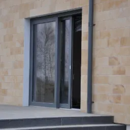 Szare, przesuwne drzwi tarasowe z dużym przeszkleniem w elewacji z jasnego piaskowca, widoczne schody wejściowe i rynna spustowa.
