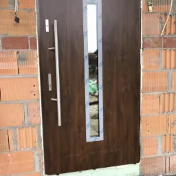 Brązowe drzwi wejściowe z długim, srebrnym uchwytem i szybą, zabezpieczoną folią, osadzone w surowym murze z czerwonej cegły, widoczne materiały izolacyjne u dołu.