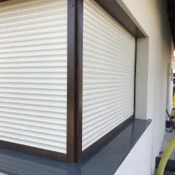 Zewnętrzny widok okna z zamontowanymi plisami w kolorze kremowym, osadzonymi w brązowej ramie, z widocznym parapetem z ciemnego granitu.