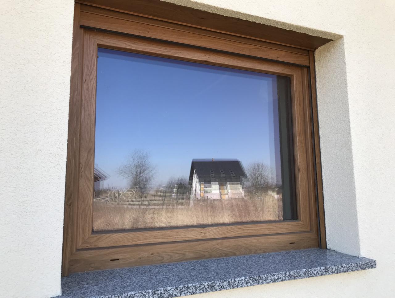 Okno z profilem w kolorze Winchester, widoczne z zewnątrz, z refleksami nieba i budynku w szybie, osadzone w białym murze na granitowym parapecie.