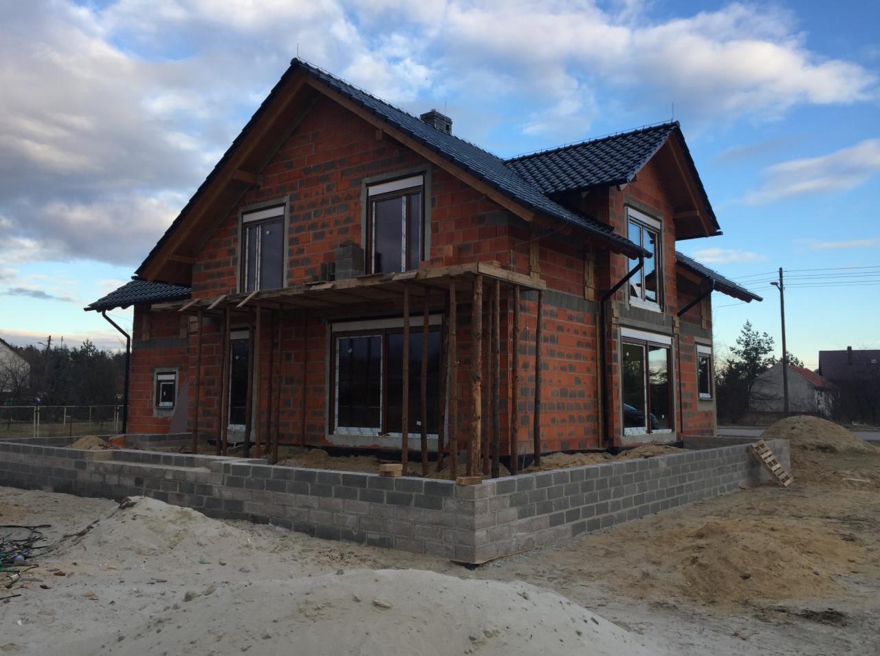 Surowy stan domu jednorodzinnego w trakcie budowy, z zamontowanymi oknami i częściowo wykonanym zadaszeniem werandy, na tle pochmurnego nieba.