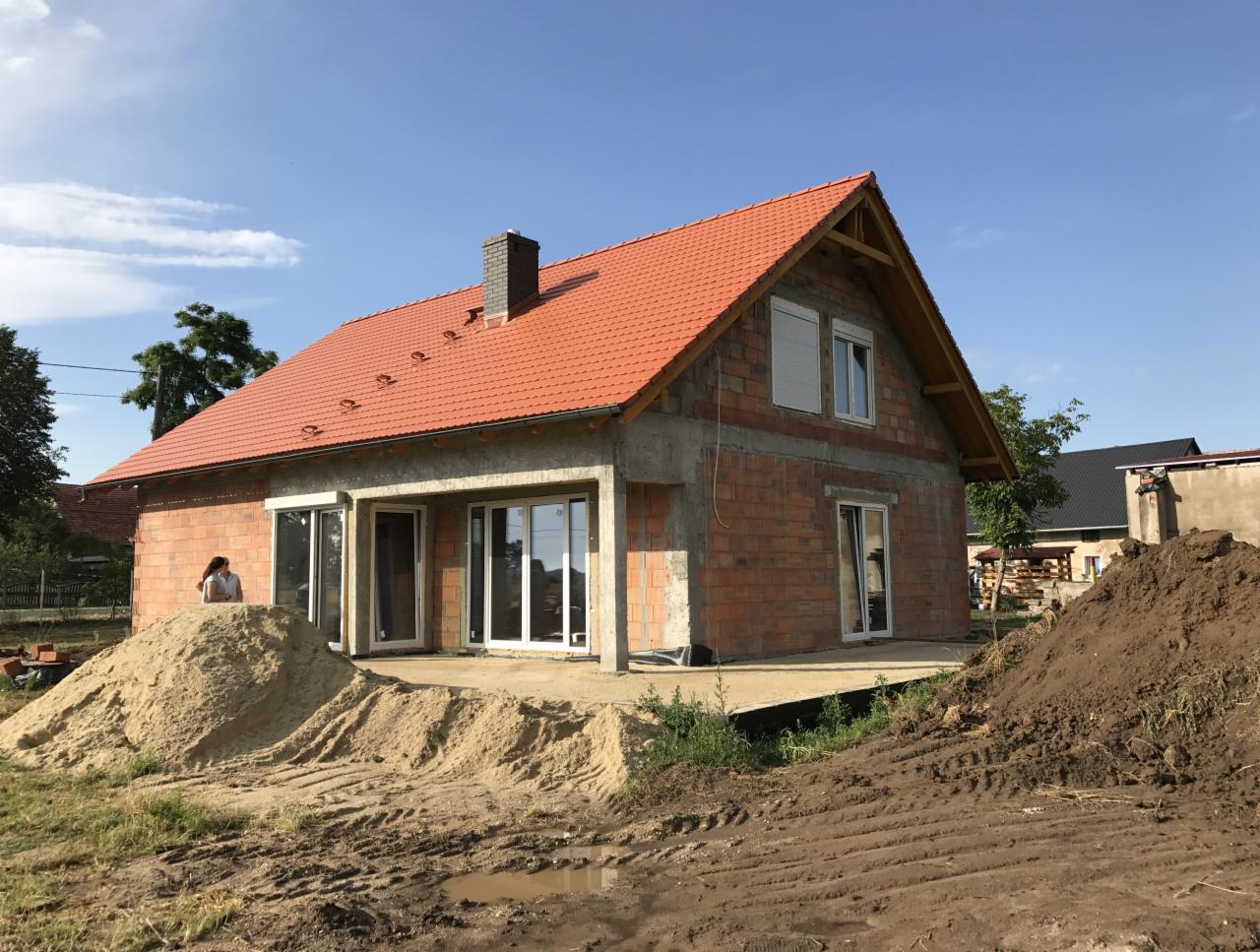 Dom w stanie surowym otwartym z zamontowanymi białymi oknami i drzwiami balkonowymi, widoczna kobieta na tle piasku, świeżo położona dachówka.