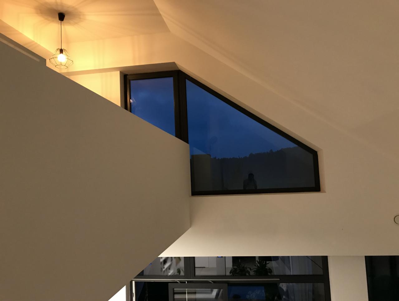 Wnętrze nowoczesnego domu we Wrocławiu z nietypowym oknem o trójkątnym kształcie, w czarnej ramie, umieszczonym pod skosem dachu, widoczne wieczorne niebo i zieleń za oknem, oświetlone ciepłym...