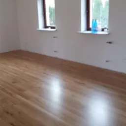 Świeżo ułożona, błyszcząca podłoga z paneli w jasnym odcieniu drewna w pokoju z dwoma oknami o łukowatych ramach. Na parapetach stoją butelki z płynem.