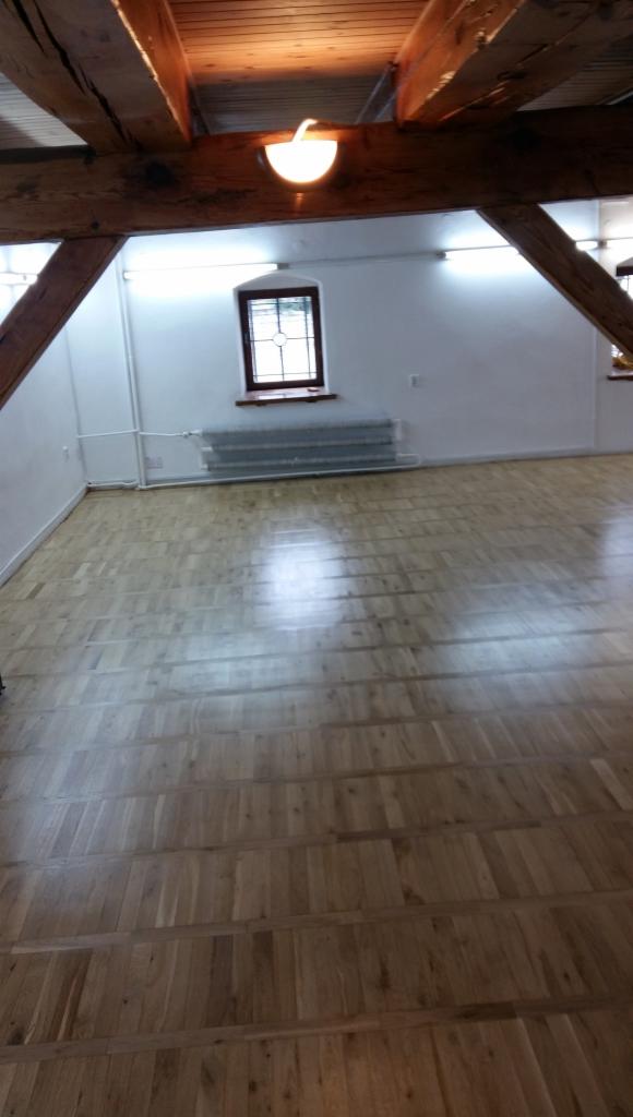 Parkiet ułożony w przestronnym pokoju z drewnianymi belkami stropowymi, widoczne okno z brązową ramą i grzejnik pod nim.