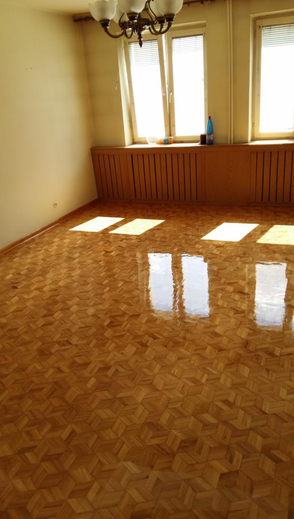 Błyszcząca, świeżo polakierowana podłoga parkietowa ułożona we wzór jodełki, z widocznymi refleksami światła słonecznego wpadającego przez okna.