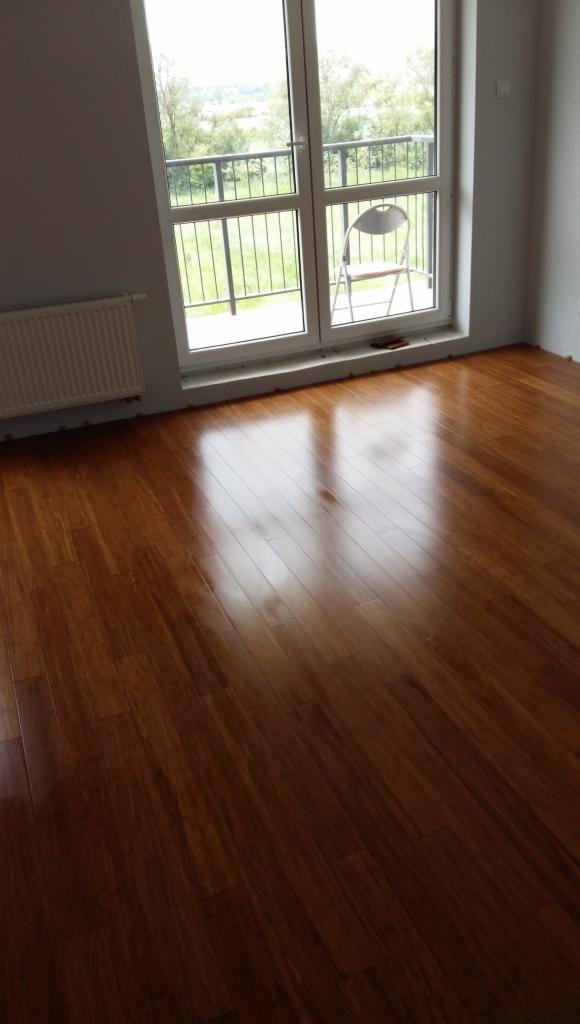 Błyszcząca, świeżo położona podłoga z ciemnego drewna w pustym pokoju z oknem balkonowym i krzesłem na balkonie.