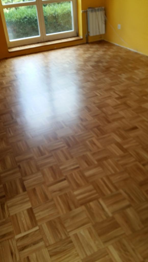 Parkiet ułożony w geometryczny wzór kwadratów, odbijający światło z okna w pokoju z żółtymi ścianami.