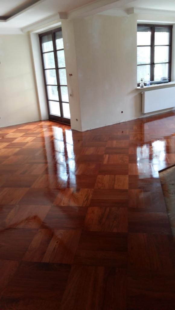 Wypolerowana, błyszcząca podłoga parkietowa ułożona w kwadraty z ciemnego drewna, odbijająca światło z okien w jasnym, pustym pokoju.