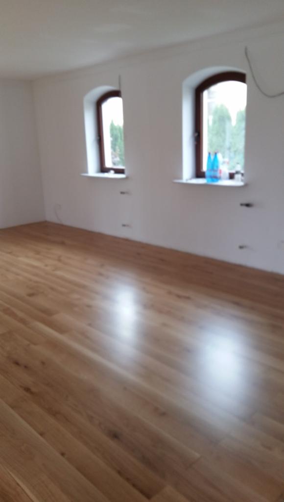 Świeżo ułożona, błyszcząca podłoga z paneli w jasnym odcieniu drewna w pokoju z dwoma oknami o łukowatych ramach. Na parapetach stoją butelki z płynem.