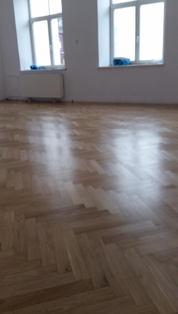 Parkiet ułożony w jodełkę francuską, widok na wykończoną podłogę w jasnym pomieszczeniu z dwoma oknami i grzejnikiem.