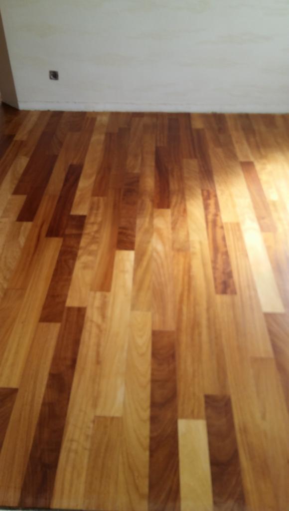 Ułożona podłoga z parkietu o zróżnicowanym odcieniu drewna, widoczne połączenia klepek i fragment ściany z gniazdkiem elektrycznym.