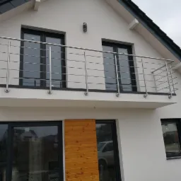 Balkon z nowoczesną, stalową balustradą na tle białej fasady domu z czarnymi oknami i drewnianą okładziną.