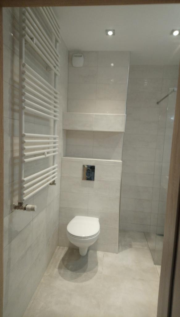 Jasna łazienka z podwieszaną toaletą, grzejnikiem drabinkowym i kabiną prysznicową w tle, wykończona płytkami w odcieniach szarości.