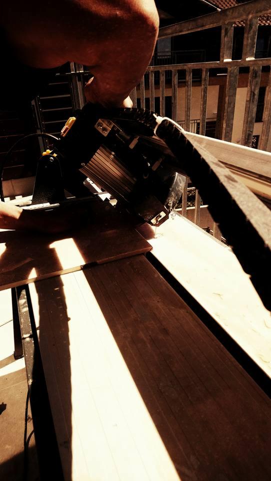 Przyciemnione ujęcie z góry: elektryczna piła ukośnica tnie drewnianą deskę na balkonie z metalową balustradą w tle.