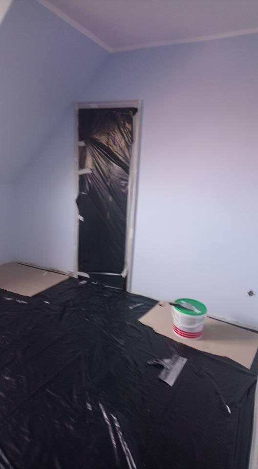 Pokój w trakcie przygotowania do malowania: ściany w jasnym odcieniu fioletu, zabezpieczone folią drzwi, podłoga zakryta czarną folią ochronną, wiadro z farbą i szpachelka na podłodze.