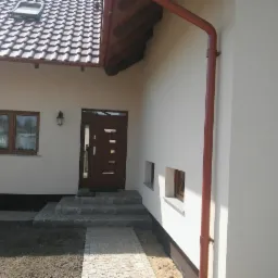 Wykończona elewacja domu jednorodzinnego z brązowymi oknami, drzwiami wejściowymi z przeszkleniami i brązową rynną, widoczne schody wejściowe z kamienia i fragment chodnika z kostki.