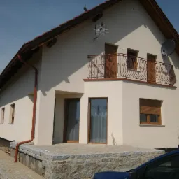 Jasny dom jednorodzinny z balkonem, brązową stolarką okienną i rynnami, widok z niskiej perspektywy, fragment niebieskiego samochodu w kadrze.