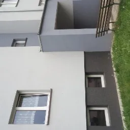 Fragment elewacji budynku wielorodzinnego z widocznym ociepleniem w kolorze grafitowym i tynkiem w odcieniach szarości, z otwartym oknem z firanką i ławką parkową w tle.