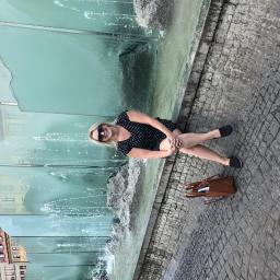 DIROM-BUD - Kobieta w okularach przeciwsłonecznych siedzi przy fontannie z przezroczystego szkła na tle miejskiej zabudowy z czerwonymi dachami.