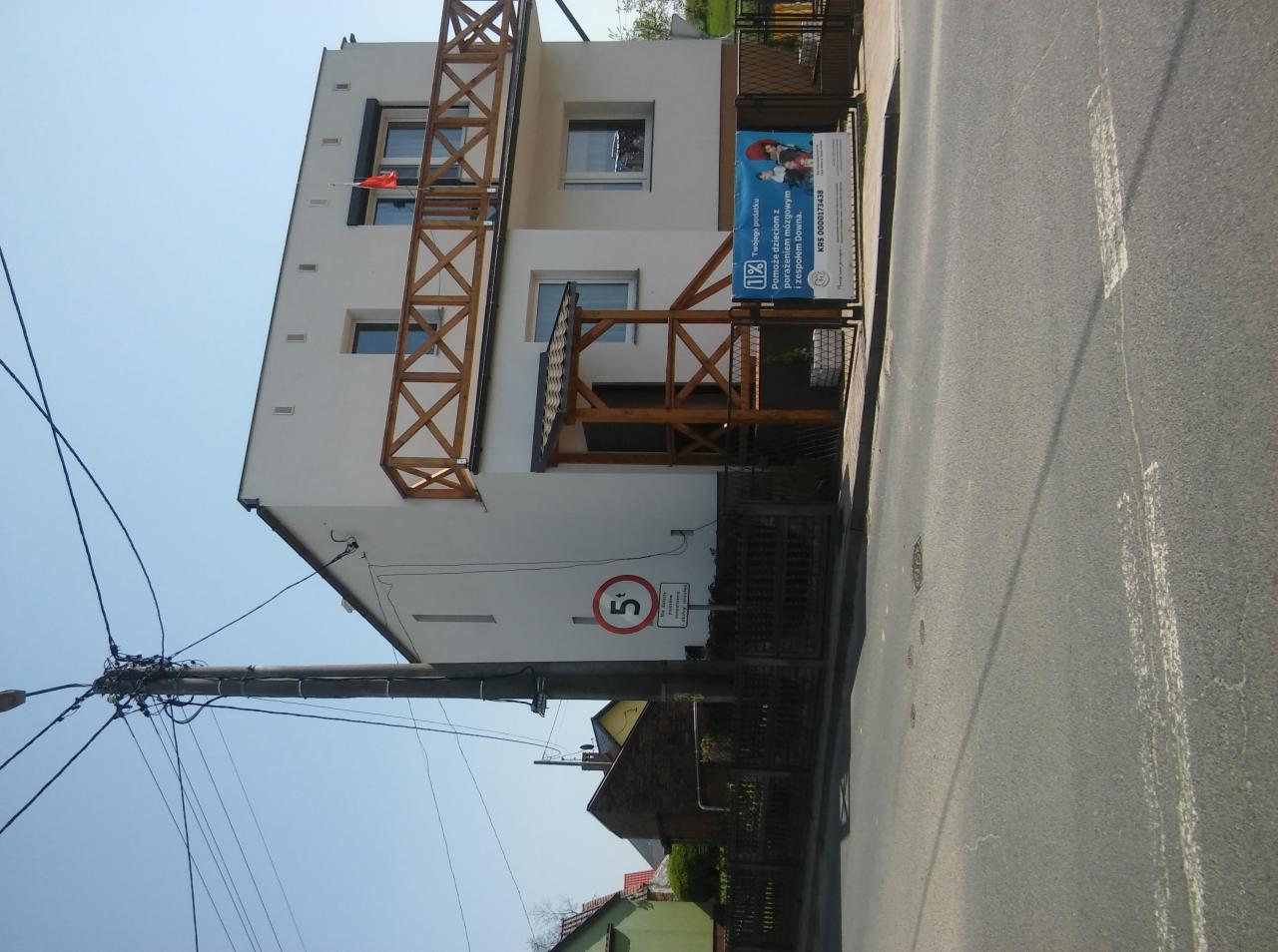 Biały dom murowany z drewnianymi elementami balkonu i zadaszenia wejścia, widoczny z perspektywy ulicy z znakiem ograniczenia prędkości i billboardem.